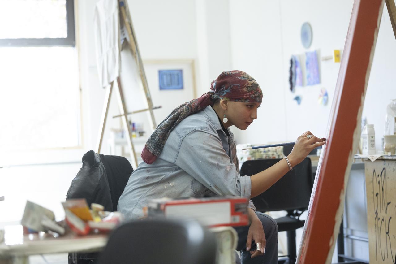 Painting Studio An art student wearing a patterned headscarf and denim shirt sits focused at an easel, carefully painting on a large canvas in a bright studio space.