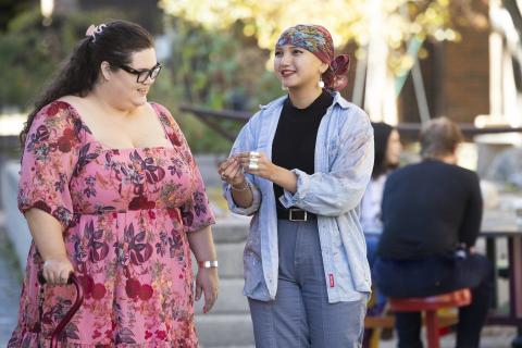 photo: Ramsey Kunkel, 2024 An outdoor scene featuring two individuals engaged in a pleasant conversation. One person wears a vibrant pink floral dress and carries a walking aid, while the other sports a colorful headscarf, a paint-splattered denim shirt, and casual pants. The background shows a sunlit garden with blurred figures at a table, creating a warm, community-oriented atmosphere.