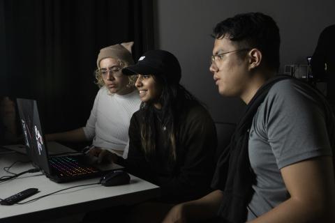 Three AUArts students collaborating at a laptop with a rainbow-lit keyboard in a dark classroom or studio.