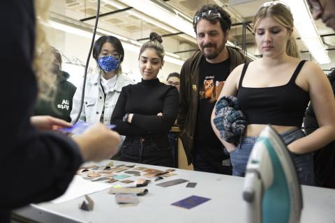 Group of AUArts students and an instructor gathered around a table observing samples of textured and colored metal pieces.
