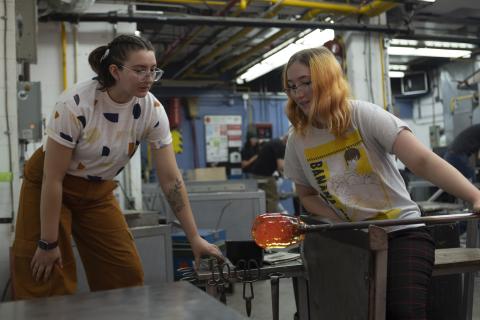 Two students shaping molten glass on a blowpipe in the AUArts glass studio.