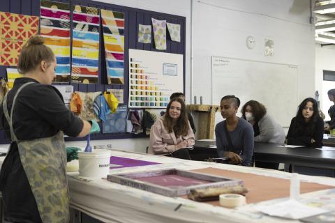 An instructor demonstrates textile printing at a large table while students watch attentively in a classroom decorated with colorful fabric samples and swatches.