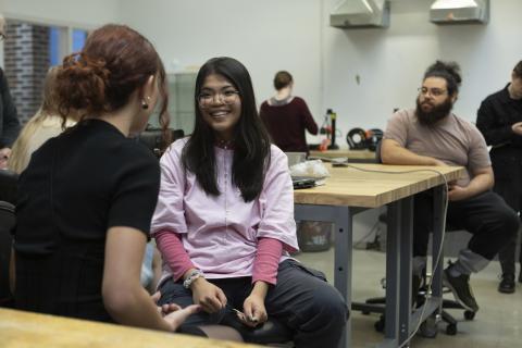 Object Design and Digital Fabrication Community Students sitting and talking around wooden workbenches in a makerspace classroom, with tools and 3D printers visible in the background.