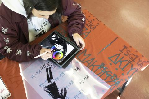 Illustration Community A student uses a stylus on an iPad to digitally illustrate a black-and-white portrait, with a color wheel open on the screen. The tablet rests on a table covered in red and pink printed sheets with bold text and graphic designs.