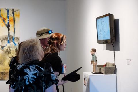 Gallery visitors view a mixed-media animation installation featuring a puppet figure on a small couch and a monitor displaying hand-drawn animation.