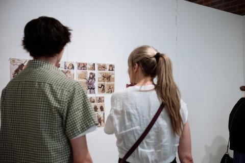 Comic Studies Community Two people view a wall display of hand-drawn comic panels featuring illustrated characters and scenes.