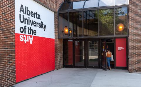 A person enters the main glass-doored entrance of Alberta University of the Arts, passing a bold red-and-white exterior wall featuring the AUArts logo.