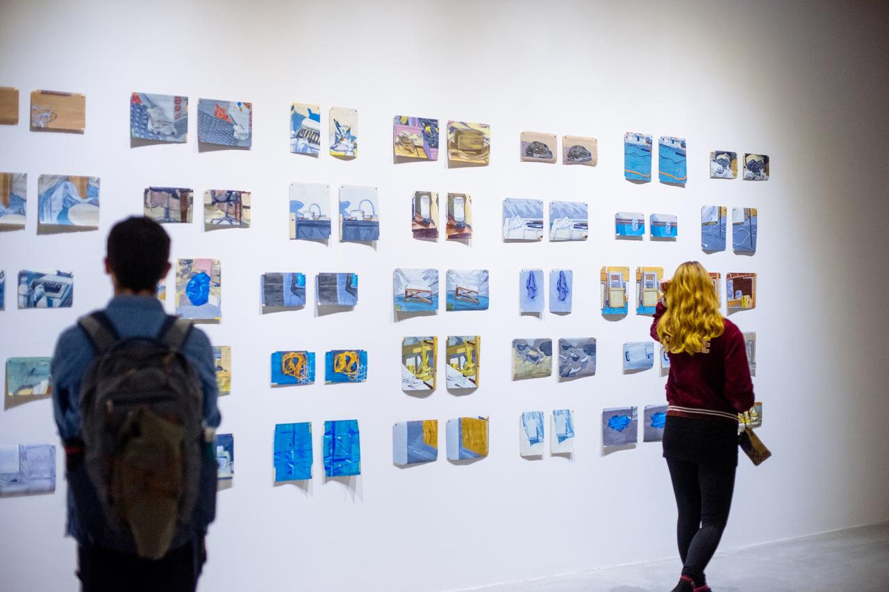Two viewers look at a grid of small paintings in blue and yellow tones