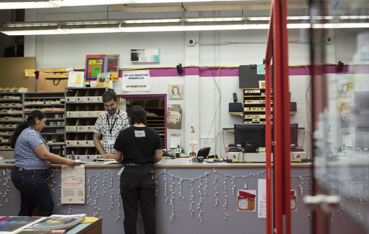 AUArts Bookstore checkout counter with two customers interacting with a staff member. The background includes shelves stocked with art supplies, a computer, and colorful decorations.