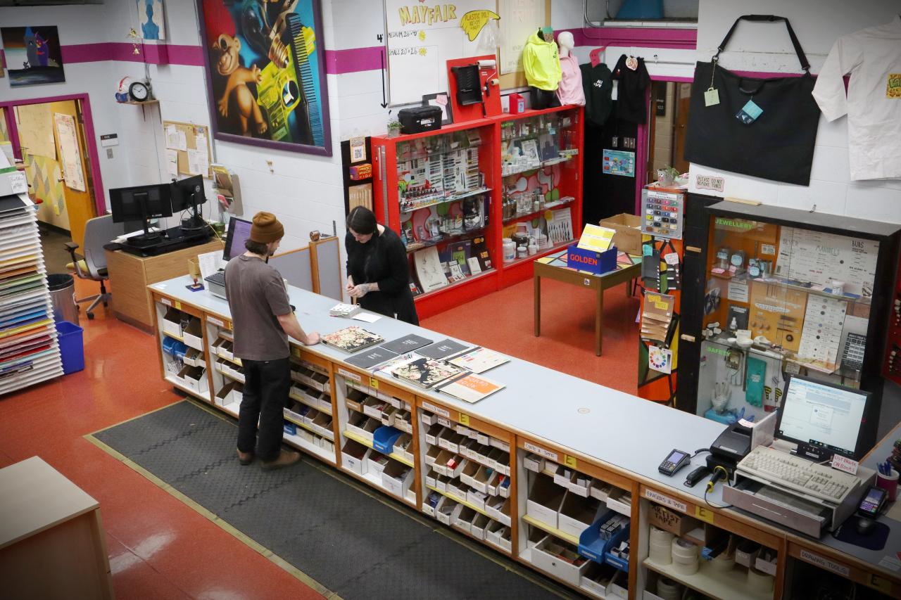 Interior view of a AUArts 'Bookstore'. An art supply store with two individuals interacting at the counter. The space features shelves stocked with various art materials, vibrant wall decor, and organized displays of products such as paper, tools, and paints. A colorful, creative environment designed for art students and enthusiasts.