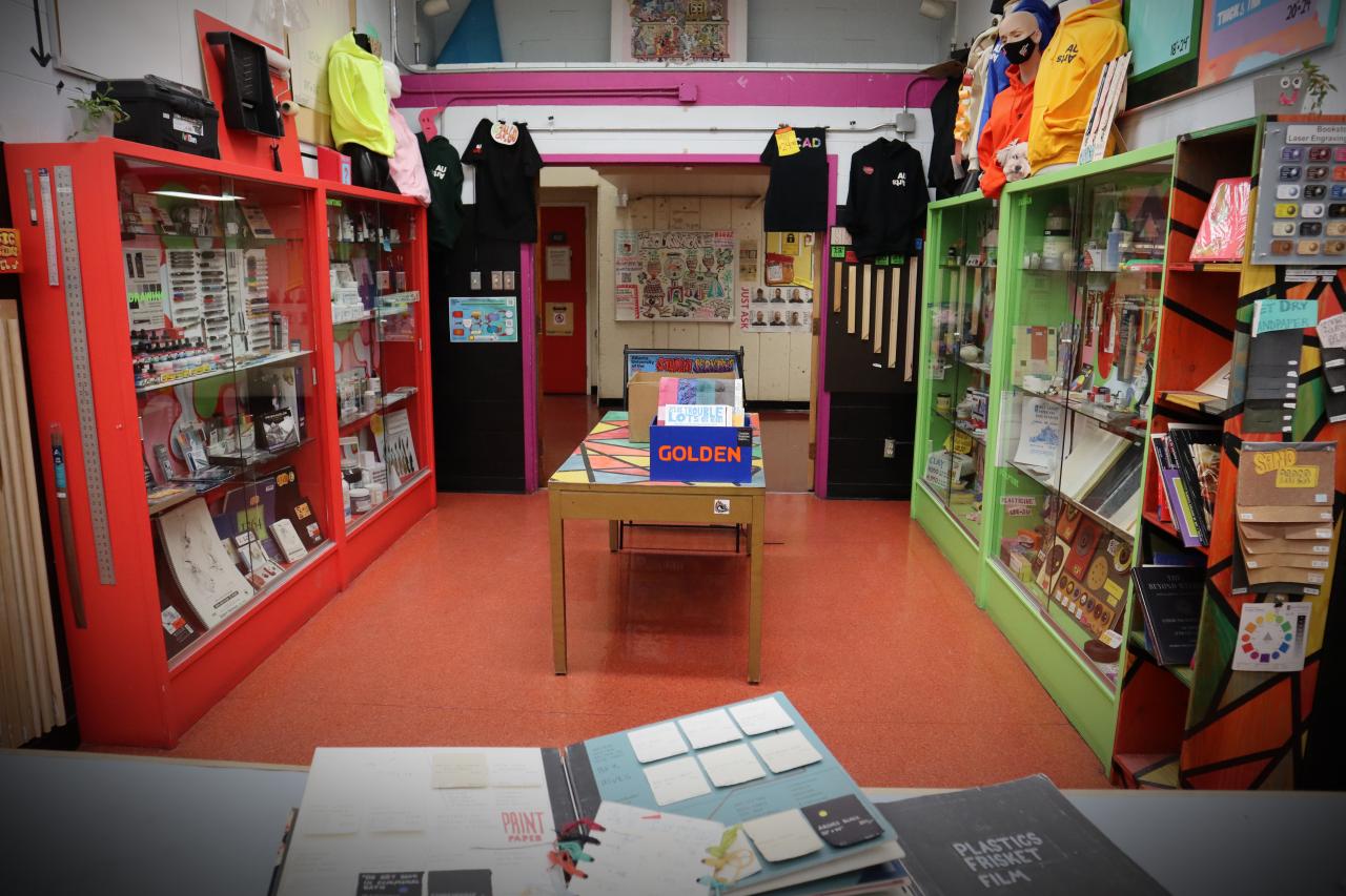 Interior of a university art supply store showcasing vibrant red and green display cases filled with art materials, tools, and merchandise. A central table holds art paper supplies, and the back wall features posters and creative displays, creating a colorful and inspiring environment for artists.