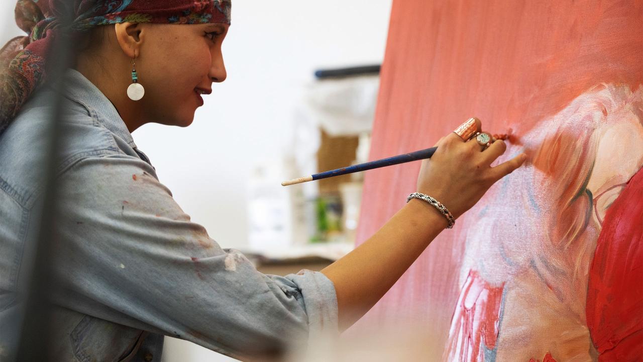 Artist painting on a large red canvas, focused on details with a paintbrush in hand. The individual, wearing a patterned headscarf and denim shirt, embodies creativity and concentration in a well-lit art studio.