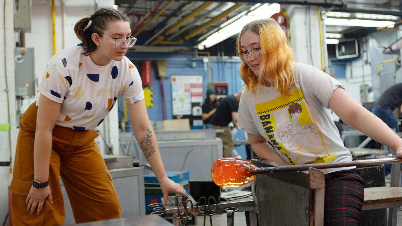 Two students collaborating in a glassblowing workshop, with one shaping molten glass on a metal pipe while the other provides guidance. The industrial studio setting features equipment, tools, and vibrant pipes in the background, highlighting hands-on artistic learning.