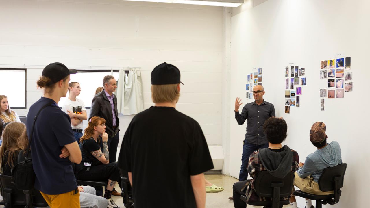 Design students engaged in a critique session, attentively listening to an instructor presenting and discussing visual research pinned on the wall. The collaborative classroom setting fosters creativity and thoughtful analysis in a dynamic learning environment.
