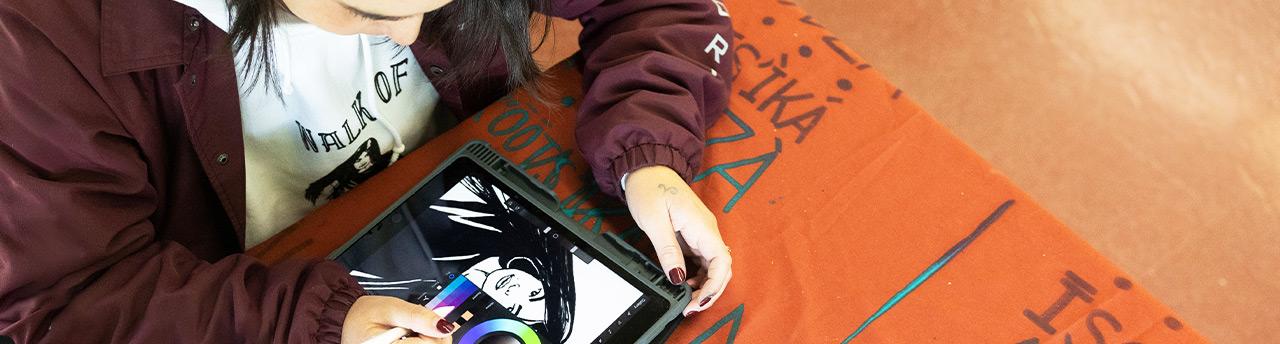 "Student working on a digital illustration using a tablet, engaged in creative design work. The workspace features a graffiti-covered table, adding an expressive and artistic atmosphere to the environment.