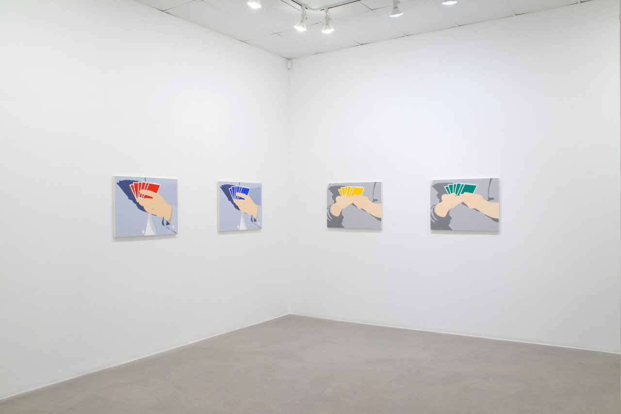 A gallery space with white walls, displaying four paintings. Each painting depicts a pair of hands holding a fan of cards in different colors: red, blue, yellow, and green. The artwork is minimalist with a limited color palette.
