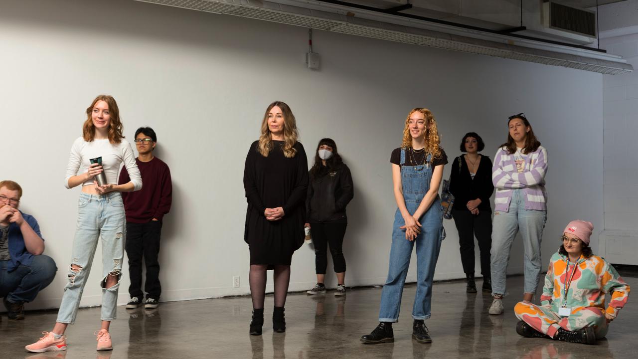 Students gathered in a contemporary gallery space, attentively listening during an art critique or presentation. The diverse group, dressed in casual and expressive styles, reflects a creative and collaborative learning environment at Alberta University of the Arts.