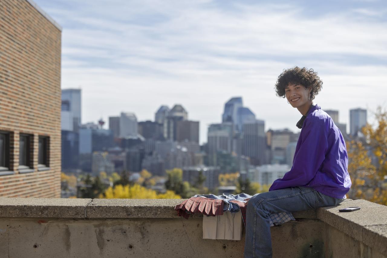 A smiling student in a purple jacket sits on a concrete ledge with the Calgary skyline in the background, surrounded by fall foliage and art materials.
