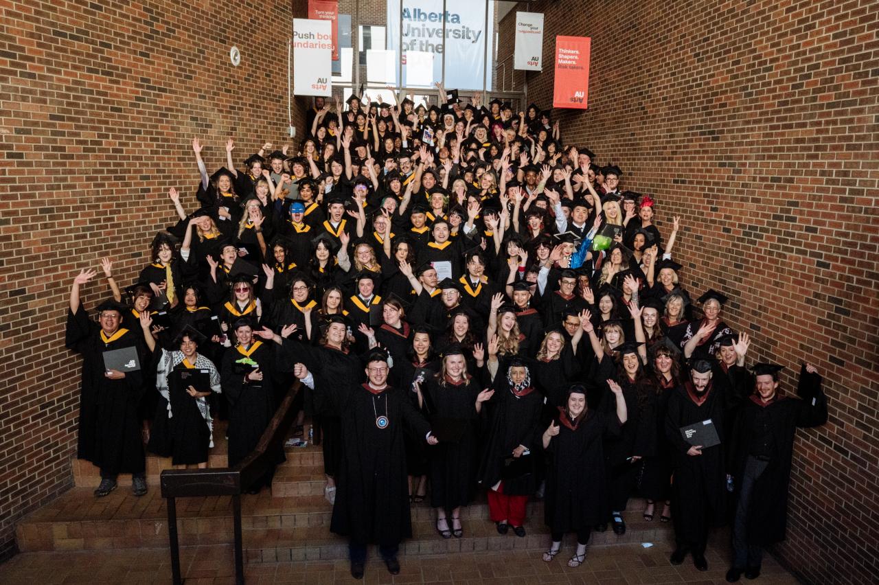 A large group of Alberta University of the Arts graduates and faculty pose together on an interior staircase, wearing black graduation gowns and caps. Many are smiling, cheering, and waving their hands in celebration. Banners above them display AUArts branding with slogans like "Push boundaries" and "Thinkers. Shakers. Makers. Risk takers." The background features brick walls and natural lighting from upper windows.