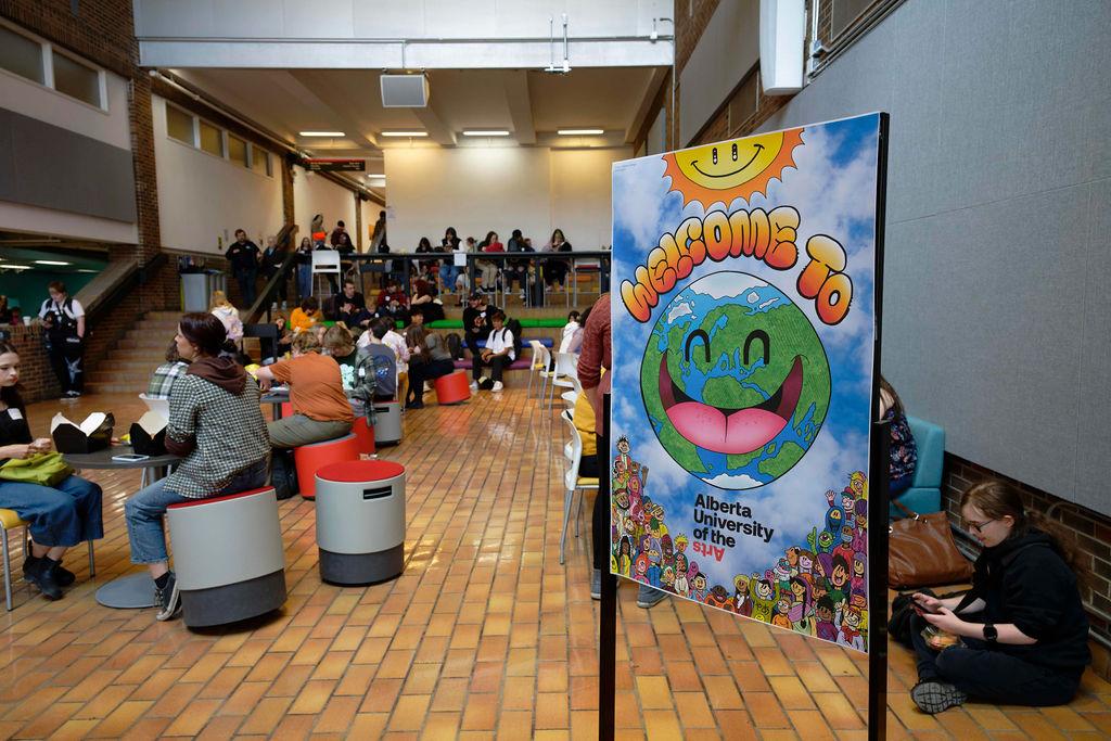 Students gather in a bright atrium space at Alberta University of the Arts, sitting at round stools and tables while socializing. A large colorful welcome sign in the foreground shows a smiling cartoon Earth under a sun with the words “Welcome To” above it and “Alberta University of the Arts” below, surrounded by illustrated characters.
