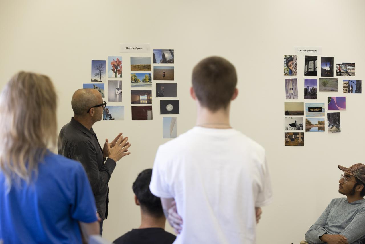 Instructor leads a critique as students view photo collages on a classroom wall labeled “Negative Space” and “Balancing Elements.”