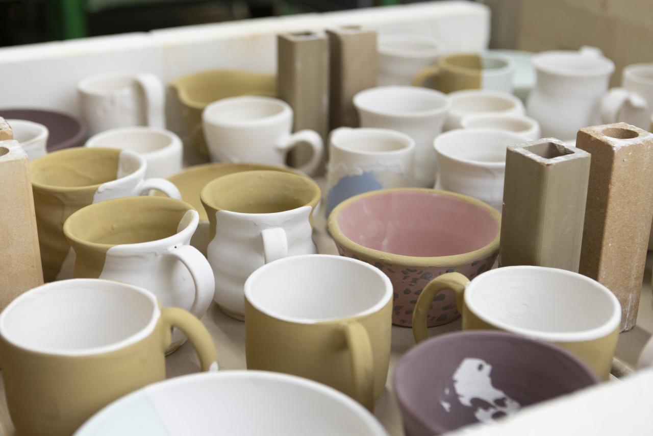 A collection of ceramic mugs and bowls in various stages of glazing, arranged together in the AUArts ceramics studio.