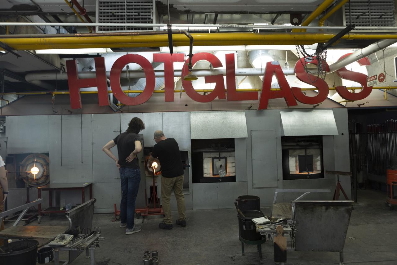 Two people working with furnaces in the AUArts glass studio under a large red “HOT GLASS” sign.