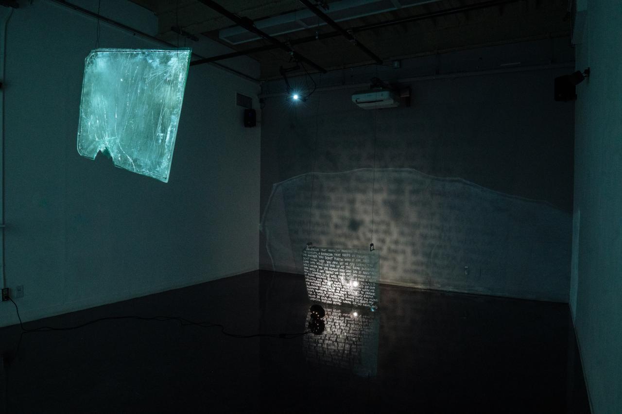 Art installation by Meghana Iyer featuring suspended broken windshields with painted text, illuminated to cast glowing reflections and shadows on the gallery wall and floor.