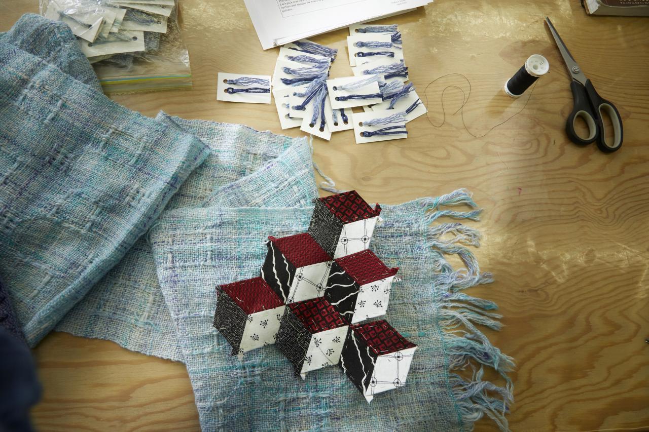 Fabric swatches, thread, and scissors on a worktable with a geometric red, black, and white textile piece placed on woven blue fabric.