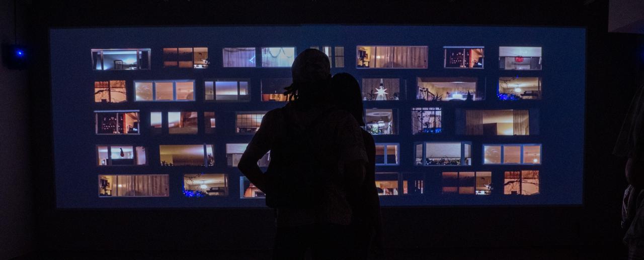 Silhouettes of two people standing in front of a large video projection of illuminated windows in a dark gallery space.