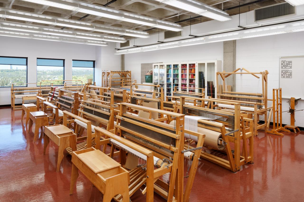 AUArts loom room with rows of wooden floor looms, stools, and shelves of colourful yarn.
