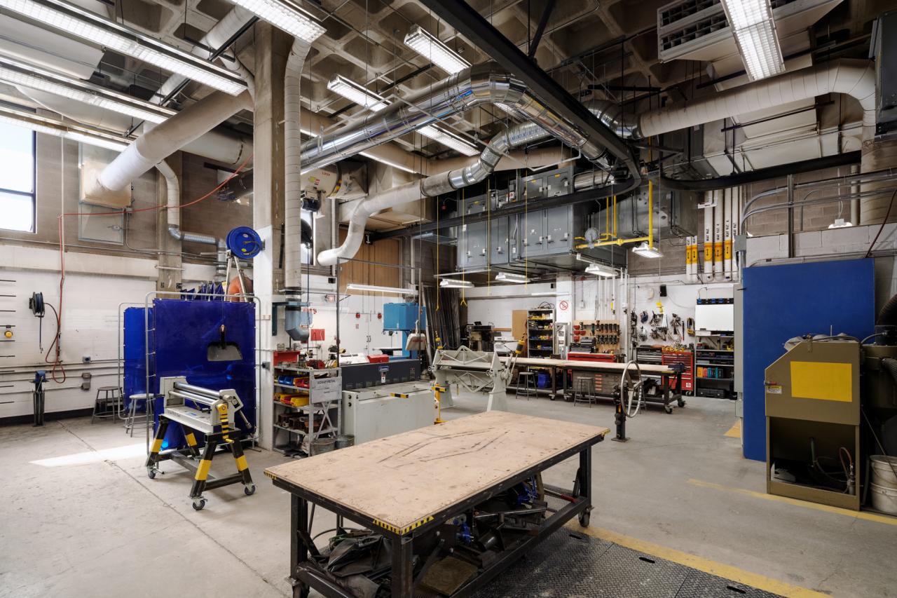 Wide view of AUArts metal shop with work tables, welding stations, ventilation systems, and industrial equipment.