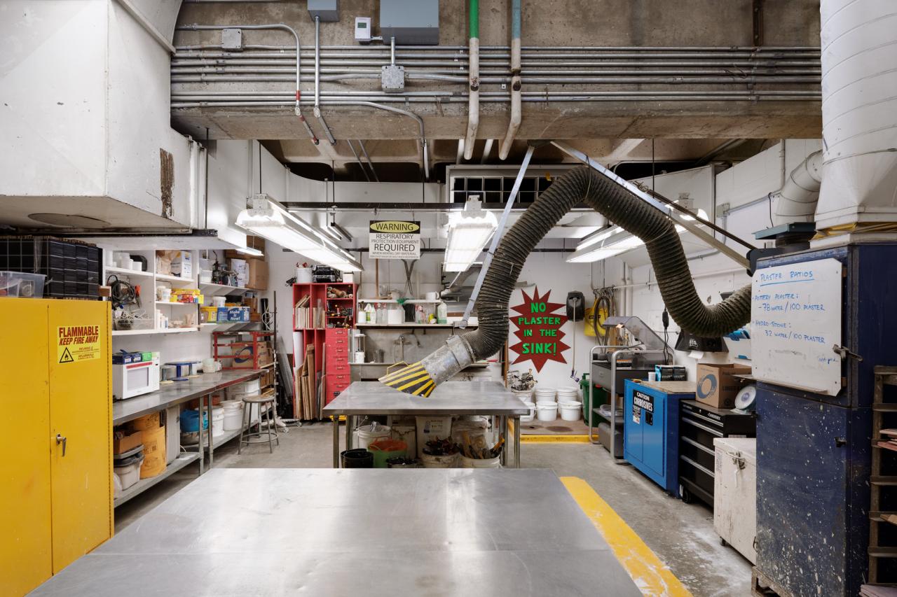 AUArts metal shop work area with large ventilation hose, stainless steel worktables, and shelves stocked with tools and supplies.