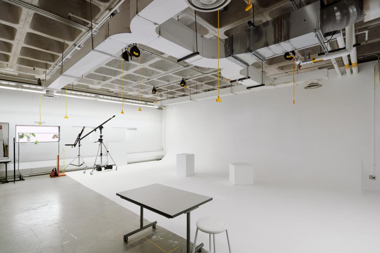A bright white photo studio with seamless white walls and floor, overhead lighting, and equipment including tripods, a crane arm, and stools.