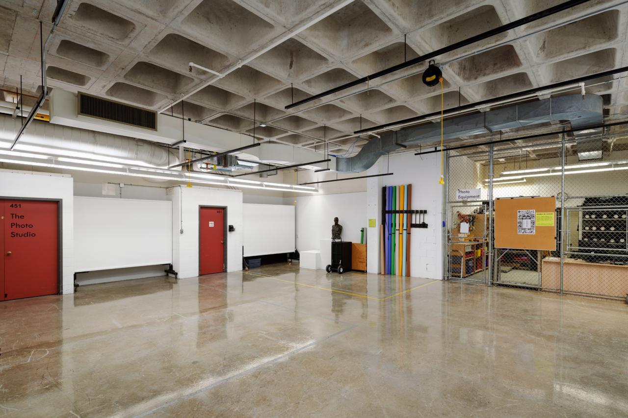 A large photo studio with red doors, white backdrops, colorful rolls of paper, and a fenced equipment checkout area with shelves and tools.