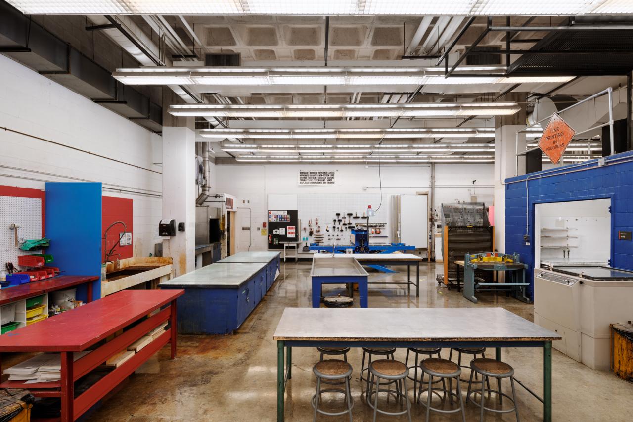 Large printmaking studio with worktables, presses, and storage cabinets, featuring bright overhead lighting and a polished concrete floor.