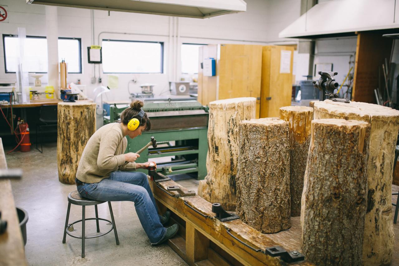 Student wearing ear protection hammering metal on a stump in the AUArts jewelry and metals studio.