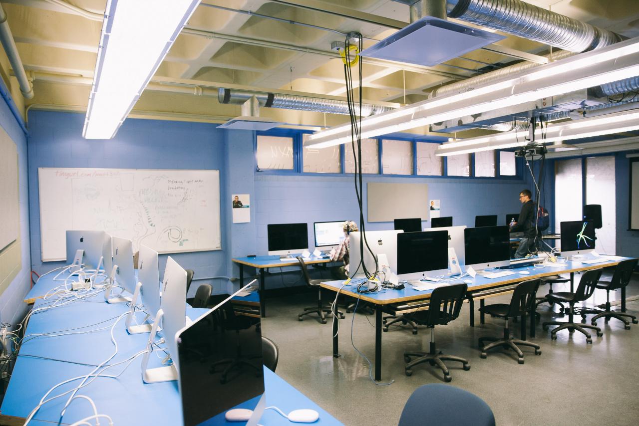 Graphic Design Mac Lab A computer lab with rows of iMacs on blue desks, chairs neatly arranged, and cables running across the tables. A whiteboard with notes and diagrams is visible on the back wall, and a person works at one of the computers while another stands nearby. The room is lit with bright overhead fluorescent lights and exposed ductwork.