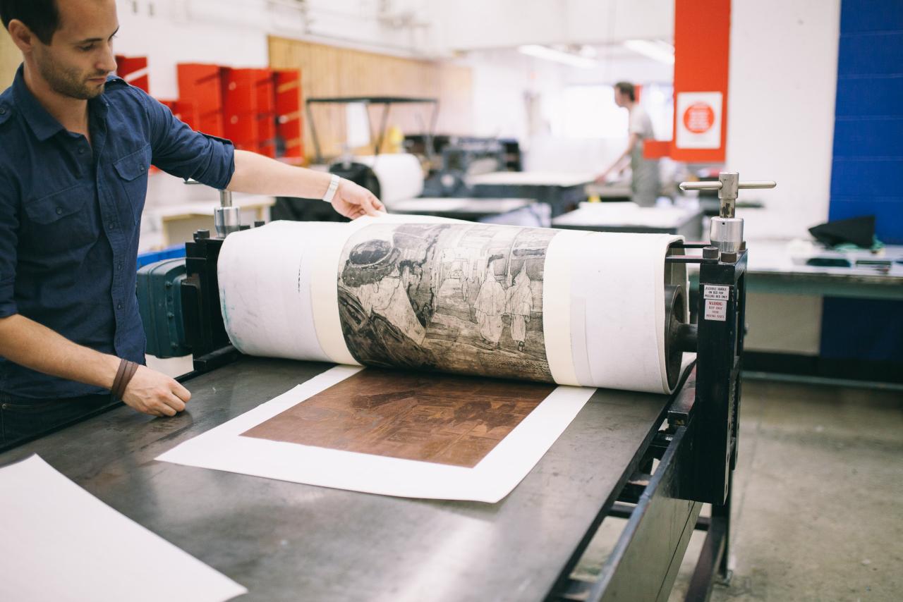 A person operates a large printmaking press, rolling out a detailed black-and-white print on paper in a studio setting.