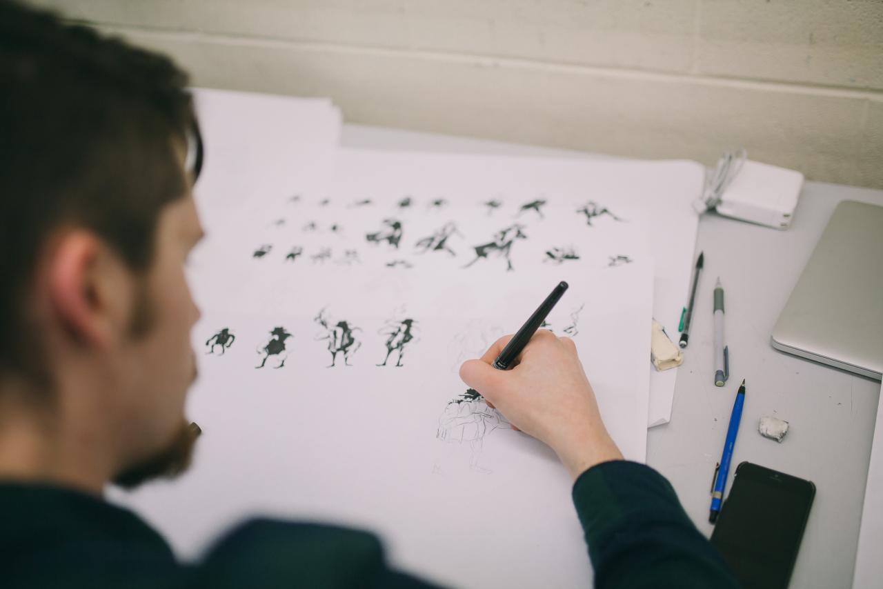 A student sketches character designs in black ink on large sheets of paper, surrounded by pens, pencils, and a laptop on a desk.