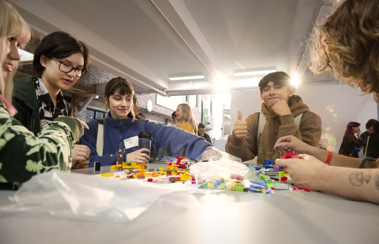 A group of AUArts students sit around a table covered with colorful LEGO bricks, smiling and building together; one student gives a thumbs-up to the camera.