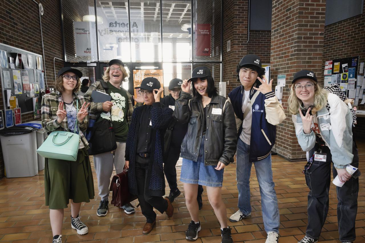 A group of smiling AUArts students pose together in the campus main mall, wearing matching black AUArts caps and making playful hand gestures during Orientation Day.