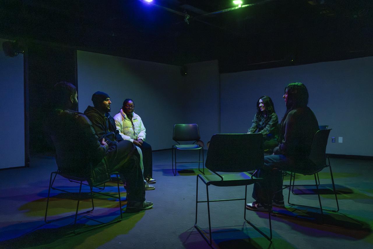 Small group seated in a circle under coloured stage lighting, engaged in discussion within a dark studio or performance space.