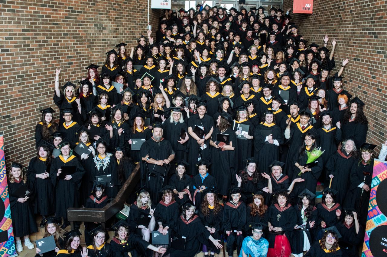 Group photo of AUArts graduates gathered on a staircase and hallway during convocation, wearing black gowns and caps with gold or red trim. Many graduates are smiling, waving, holding diploma folders, flowers, or celebrating with classmates beneath AUArts banners and colourful “100” centennial signage.