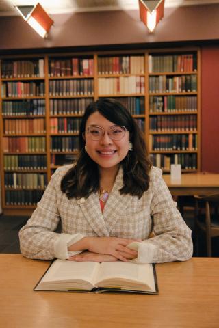 A person sits at a wooden table in a library, smiling at the camera with an open book in front of them and shelves of books lining the background.