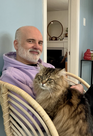 A person in a lavender hoodie sits in a wicker chair, smiling while holding a large, fluffy brown tabby cat with green eyes. The cat rests calmly in their lap. In the background, a softly lit room features a fireplace mantle with a round mirror and small decorative objects.