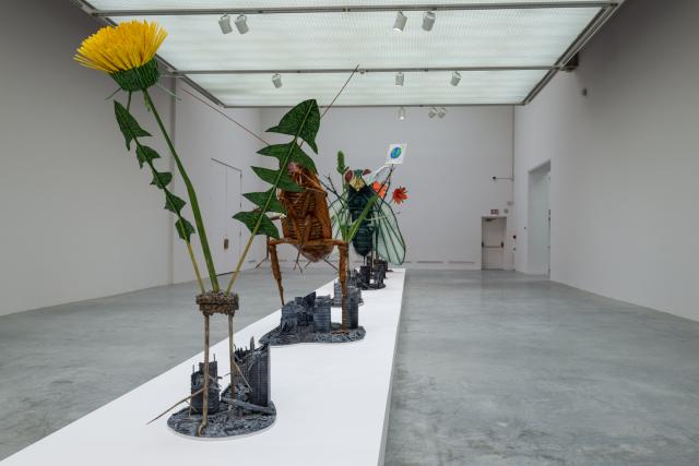 side view - long platform with larger than life sculptures of a dandelion, cockroach, barnyard grass, scarlet pimpernel and gypsy moth