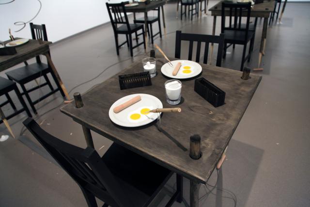 View of a table. Two plates with two eggs on each place, a hotdog is on the plate. There is a wooden stake pointing at the plate. There are two wooden cages holding a hot dog each. There are two glasses of milk at different volumes, each has a milky tongue reaching from the milk. 