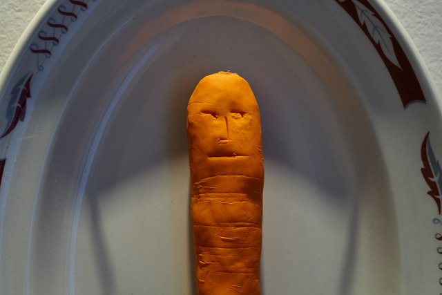 Detail of a carrot hanging on the wall. It has a solemn face mimicking the lines of the carrot. 
