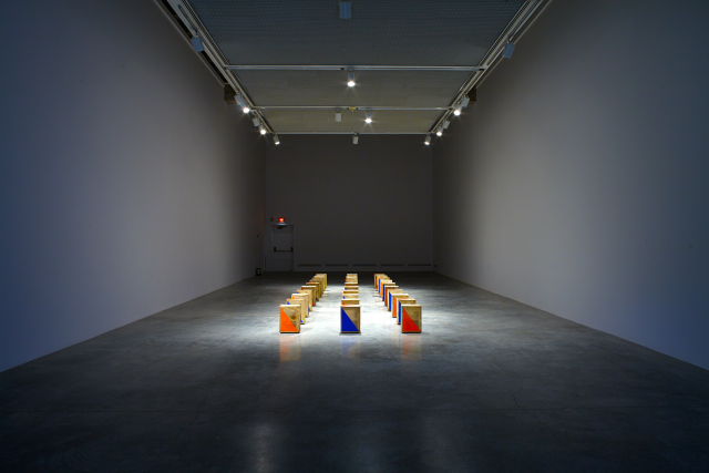 Distanced shot of the gallery space with the objects lined up, rows of three by eight. 